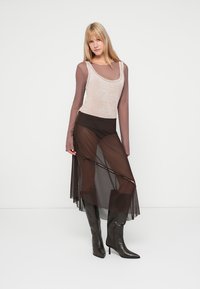 Viacvrstvový outfit s svetlou béžovou bezrukávovou blúzkou, dlhou hnedou priesvitnou sukňou s gombíkovými detailami a čiernymi čižmami po kolená.
