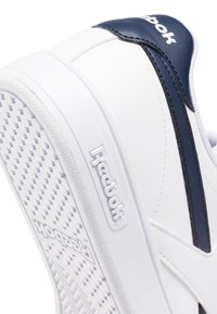 Baskets blanches avec accents navy, en matériau synthétique lisse, semelle texturée et logo surélevé sur le côté. Design épuré et contemporain.