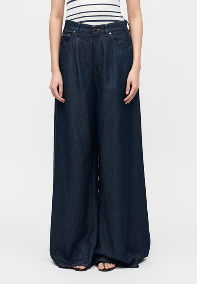 Wide-leg dark denim jeans worn with striped top, hands relaxed at sides, feet in black sandals visible under hem.