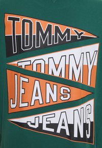Zelené tričko s velkým, tučným textem: "TOMMY" a "JEANS" v černé, bílé a oranžové výřezech geometrických tvarů, s hladkým povrchem.