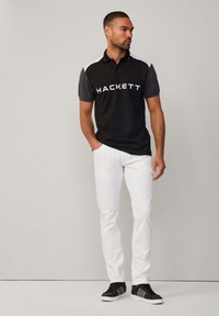 Polo negro con mangas grises, que presenta acentos de color rojo en el cuello y el logo "HACKETT" en blanco. Combinado con pantalones blancos y zapatillas negras.