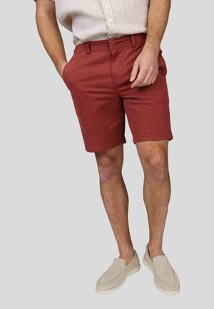 Shorts rouges en coton, dotés d'un devant plat et de poches latérales. Associés à des chaussures slip-on beige clair, sur un fond gris neutre.