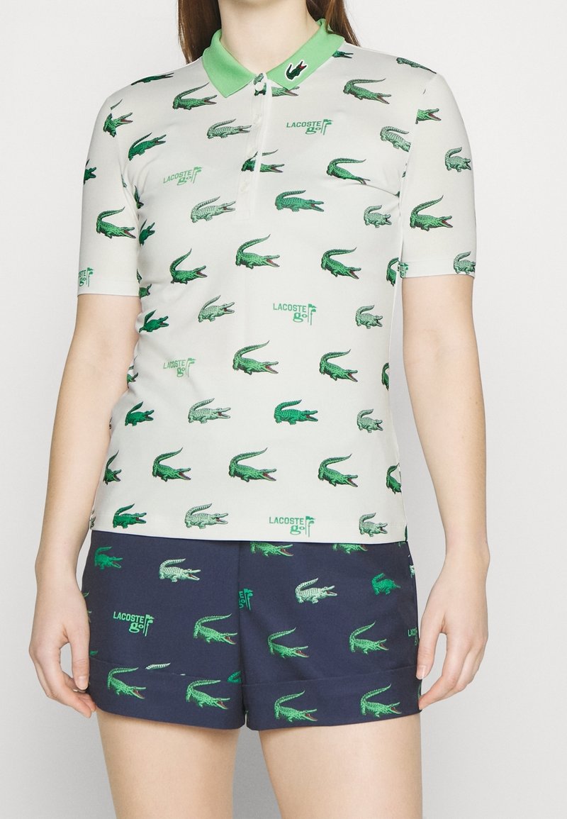 Krátký rukáv polokošile v bílé barvě se zelenými vzory krokodýlů, se zeleným límcem a nápisem "Lacoste golf". Hladká textura.