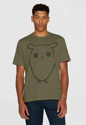 Giovane uomo con t-shirt verde oliva con grande contorno nero di un gufo stilizzato sul davanti, in piedi davanti a uno sfondo chiaro e semplice.
