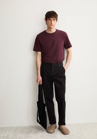 Bordeauxkleurig T-shirt met korte mouwen, zwarte broek met zakken, beige instapschoenen en een zwarte tote bag met een wit logo. Neutrale achtergrond.