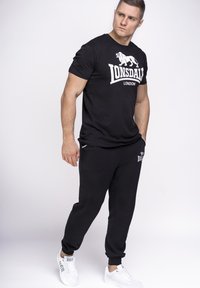 Fekete Lonsdale póló fehér logóval, fekete jogger nadrággal, melyen logo és fehér díszítések találhatók. A modellen fehér sportcipő van.
