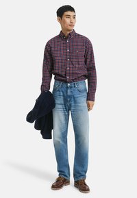 Mann in kariertem roten und blauen Hemd, hellblauen Jeans, braunen Lederschuhen, hält in seiner linken Hand eine dunkelblaue Jacke.