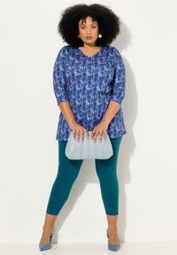 Långärmad V-ringad tunika i lila och blått mönster, matchad med teal leggings; håller en ljusblå, plisserad handväska och har på sig blå klackskor.