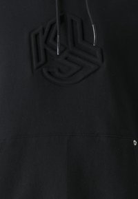 Czarna tkanina z wypukłym haftem geometrycznego logo i czarnymi sznurkami ściągającymi z metalowymi końcówkami wiszącymi powyżej.