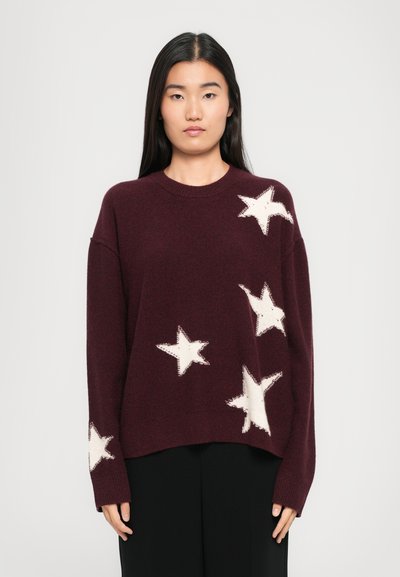 Donna con lunghi capelli scuri che indossa un ampio maglione bordeaux con motivi a stelle bianche, in piedi contro uno sfondo chiaro e uniforme.
