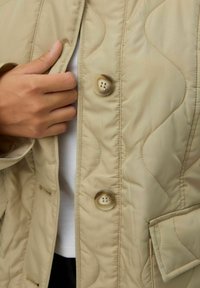 Beige quiltet jakke med en glat tekstur, der har en klassisk krave og tre knapper. En hånd ses gribe fat om lapelen.