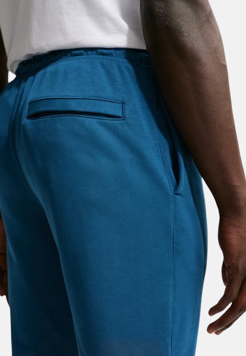 Personne portant un pantalon bleu avec des poches à l'arrière et sur les côtés, debout avec le bras droit détendu le long du corps.