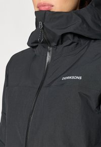 Chaqueta impermeable negra con cuello alto, cremallera frontal y tejido texturizado. Presenta un pequeño detalle de logo en el área del pecho.
