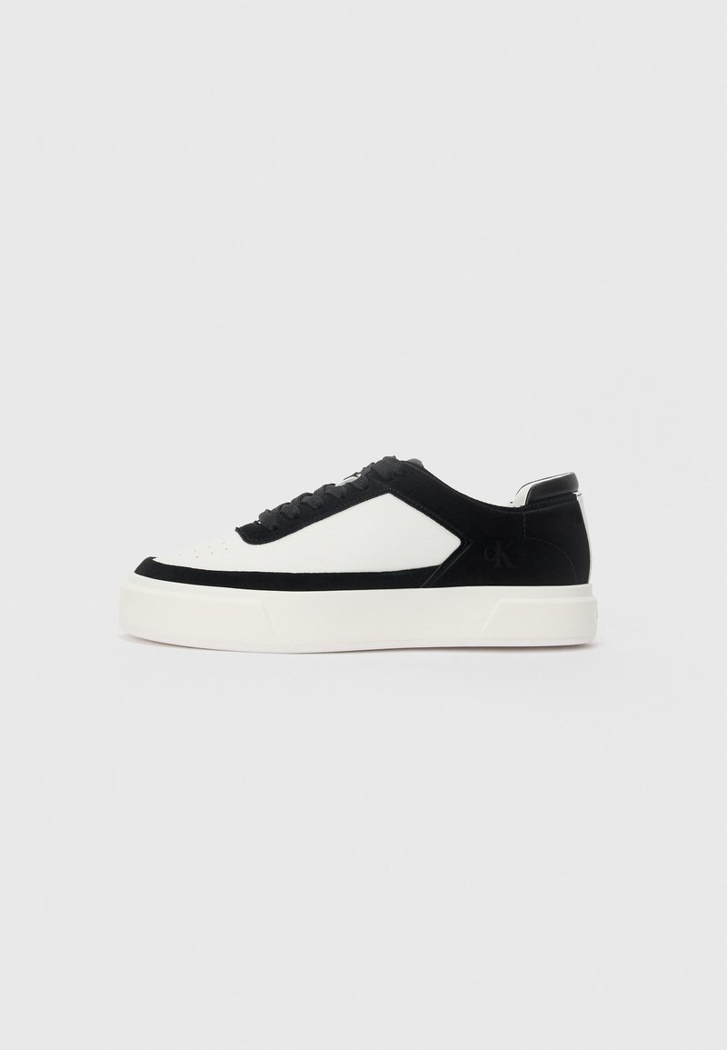 Lage sneaker van zwart suede met witte leren panelen, zwarte veters en een dikke witte rubberen zool, weergegeven vanaf de buitenzijde.