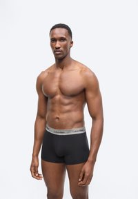 Homme musclé debout, portant un boxer Calvin Klein noir, sur un fond gris clair uni, faisant face à l'avant avec une expression neutre.