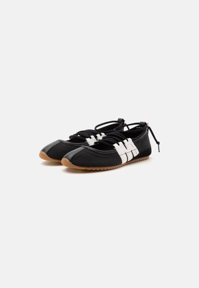 Paire de chaussons de ballet plats noirs et blancs avec semelles en caoutchouc beige et lacets autour de la cheville sur un fond clair uni.