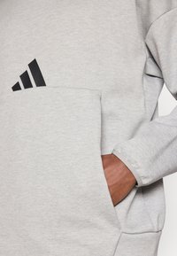 Pilkas džemperis su tekstūruotu paviršiumi, turintis didelę kišenę ir išskirtinį juodą Adidas logotipą viršutiniame kairiajame kampe.