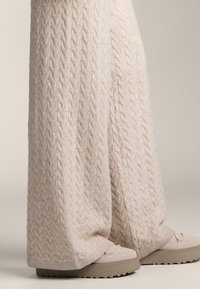 Široké, pletené nohavice krémovej farby s vrkočovým vzorom, skombinované so svetlo béžovými slip-on topánkami s hrubou textúrovanou podrážkou.