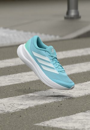 Chaussure de course Adidas bleu clair avec des rayures blanches flottant au-dessus d'un passage piéton texturé sur un pavement gris.