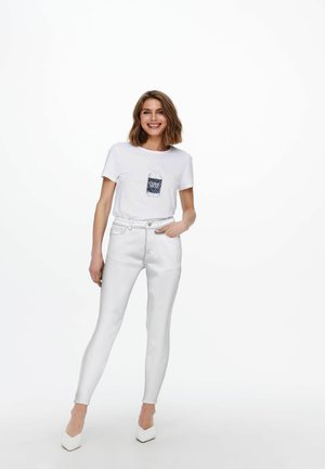 Mujer sonriente de pie, vestida con vaqueros blancos, tacones blancos y una camiseta blanca con un diseño gráfico azul.