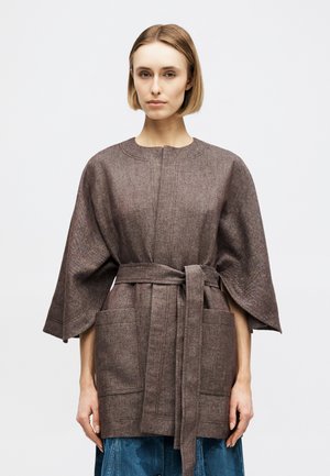 Nő rövid szőke hajjal, barna, texturált gyapjúkabátot visel övvel, széles ujjal és nagy első zsebekkel.