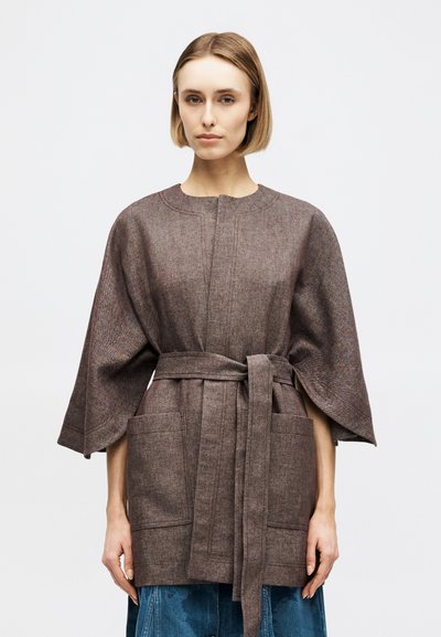 Femme aux cheveux blonds courts portant un manteau en laine marron texturé avec ceinture, manches larges et grandes poches avant.