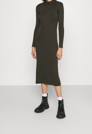 Femme portant une robe midi à manches longues côtelée de couleur vert olive, associée à des chaussettes blanches et des bottines noires, sur un fond uni.