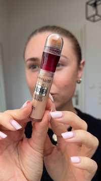Femme tenant le correcteur multi-usages instantané anti-âge de Maybelline New York avec applicateur en éponge, sous la lumière naturelle à l'intérieur.