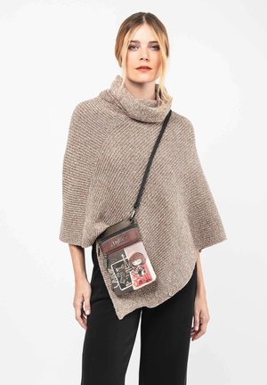 Mujer con cabello rubio que lleva un poncho de punto beige y pantalones negros, cargando un pequeño bolso cruzado con diseños de dibujos animados y texto.