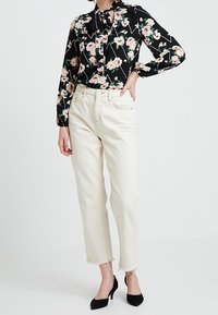 Blusa nera a fiori con maniche lunghe e collo a fiocco, abbinata a jeans beige a gamba dritta con orlo grezzo e scarpe con tacco nere.