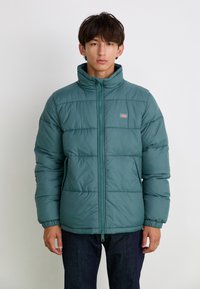 Cilvēka puffer jaka zaļā krāsā ar augstu apkakli, ar rāvējslēdzēju un ribotiem piedurknēm. Iezīmē horizontālas quiltētas zonas un mazu logo uz krūtīm.