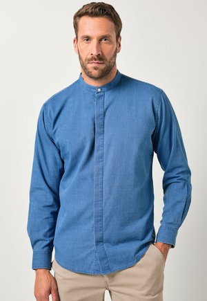 Uomo con capelli corti e barba, che indossa una camicia blu con colletto alla mandarino e pantaloni beige, in piedi con una mano in tasca.