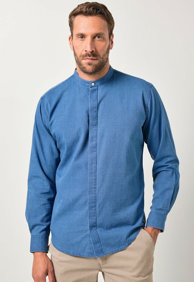 Man met kort haar en baard, gekleed in een blauw hemd met mandarin kraag en beige broek, staande met één hand in zijn zak.
