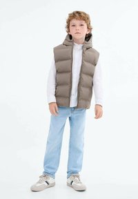 Rjav puffer telovnik s kapuco, v kombinaciji z belo srajco z gumbi, svetlo modrimi kavbojkami in sivimi supergami s temnimi črtami.