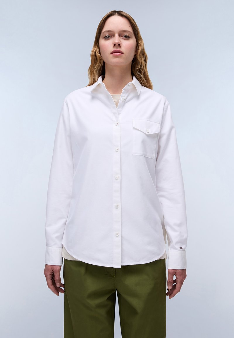 Camicia bianca con bottoni, dotata di un'unica tasca sul petto, realizzata in tessuto morbido, maniche lunghe con polsini abbottonati, abbinata a pantaloni verde oliva.