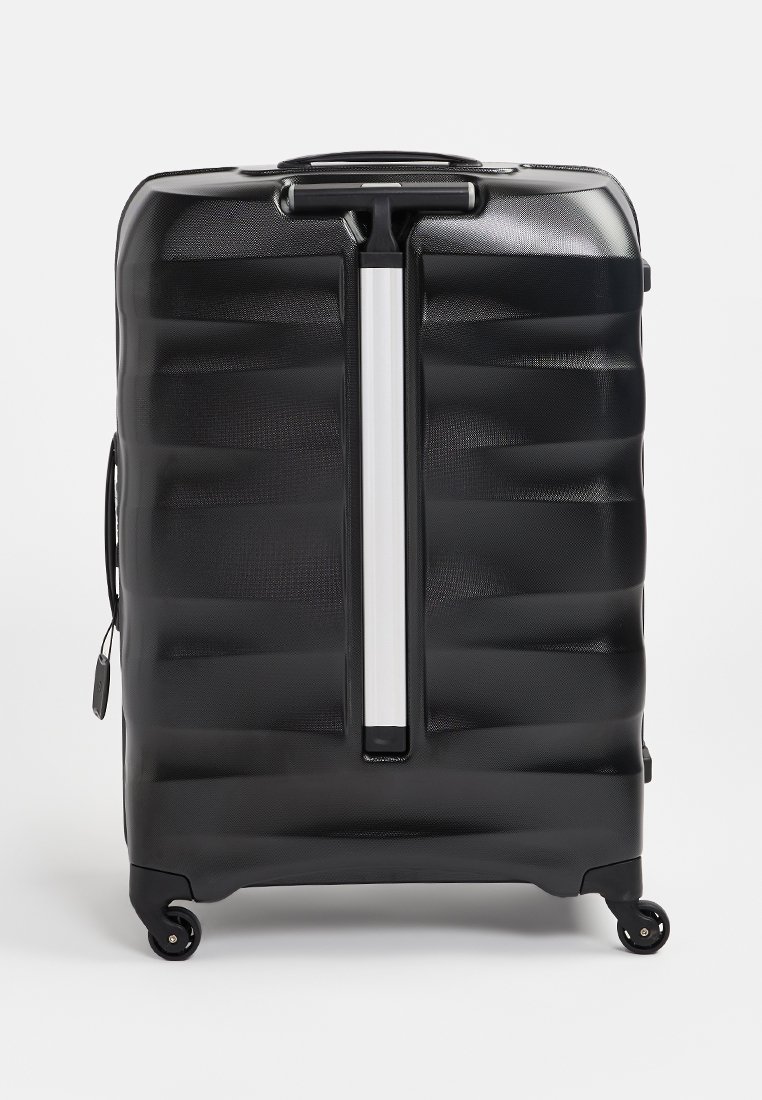 Valise rigide noire avec des rainures horizontales, poignée télescopique et quatre roues pour une mobilité aisée. Présente un accent central argenté.