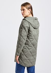 Veste en olive vert matelassée avec capuche, présentant un motif en losanges et un patch logo sur l'épaule. Manches longues et coupe mi-longue.
