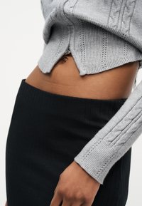 Κοντινό πλάνο στη μέση ενός ατόμου που φοράει γκρι πλεκτό κοντό πουλόβερ και ψηλόμεση μαύρη ριμπ φούστα.