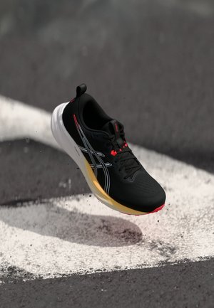 Czarny but sportowy z teksturowaną cholewką, białą i żółtą podeszwą środkową, czerwonymi akcentami i kontrastującymi czarnymi sznurowadłami, uniesiony w powietrzu nad malowanym podłożem.