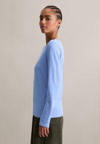 Junge Frau mit geflochtenem Haar steht seitlich, trägt ein hellblaues Langarmshirt und einen dunkelgrünen Rock vor einem einfarbigen Hintergrund.