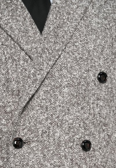 Nærbillede af en grå herringbone-mønstret dobbeltbrystet frakke med brede revers og tre synlige sorte knapper over et sort beklædningsstykke.