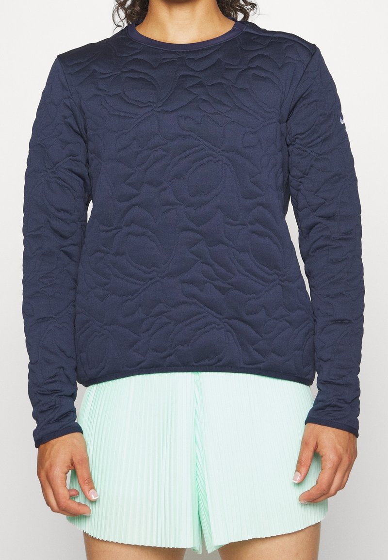 Personne portant un haut à manches longues bleu marine texturé avec un matelassage floral et un short plissé vert menthe clair, debout devant un fond uni.