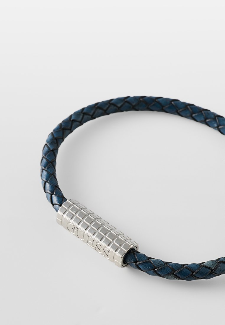Bracelet tressé en cuir bleu avec un fermoir en métal argenté texturé gravé de "GUESS". Le design est circulaire et flexible.