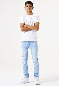 Camiseta blanca de manga corta, jeans ajustados de color azul claro y zapatillas blancas. Diseño sencillo sin patrones visibles. Fondo de estudio.