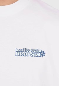 T-shirt en coton blanc avec un texte bleu indiquant "local ice dealer DROPSIZL" accompagné d'un graphique d'étoile. Design standard à col rond.