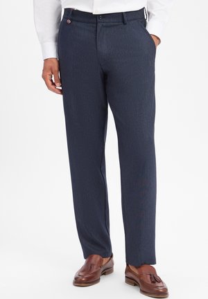 Mann trägt marineblaue Anzughose, weißes Hemd und braune Leder-Tassel-Loafer, steht mit einer Hand in der Tasche vor weißem Hintergrund.