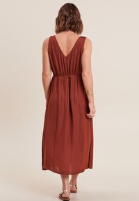 Langes, rostfarbenes Kleid mit V-Ausschnitt am Rücken, geraffter Taille und fließendem Stoff, der eine leichte Textur und ein knöchellanges Design aufweist.