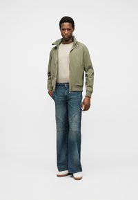 Άνδρας με ανοιχτό πράσινο μπουφάν  με φερμουάρ, μπεζ πουκάμισο, μπλε τζιν φαρδιάς γραμμής και λευκά παπούτσια, στέκεται με το ένα χέρι στην τσέπη μπροστά σε λευκό φόντο.