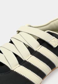 Zwarte stoffen schoen met crèmekleurige elastische veters en decoratieve crèmekleurige strepen. Heeft een gestructureerd oppervlak en gestikte accenten aan de zijkant.