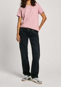 Camiseta corta de manga en color rosa con logo en relieve. Jeans negros con tachuelas metálicas a lo largo del lado, combinados con zapatillas plateadas.
